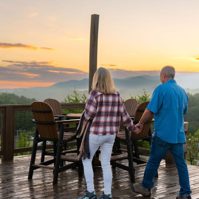 Two people holding hands on a wooden deck overlooking a scenic sunset with mountains.