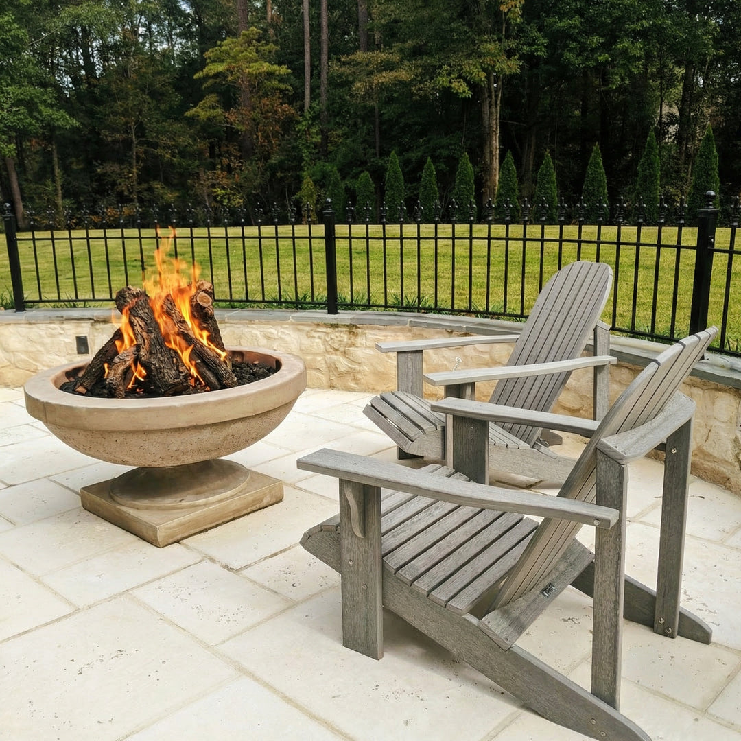 Two Adirondack chairs around a fire pit on a patio with a fence and trees in the background.