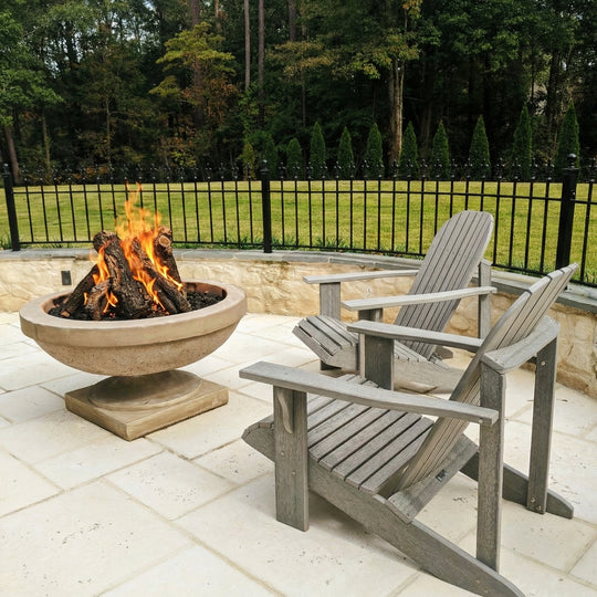 Two Adirondack chairs around a fire pit on a patio with a fence and trees in the background.
