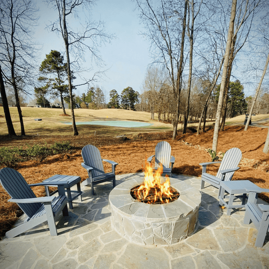 Fire pit with Adirondack chairs in a park-like setting with trees and grass.