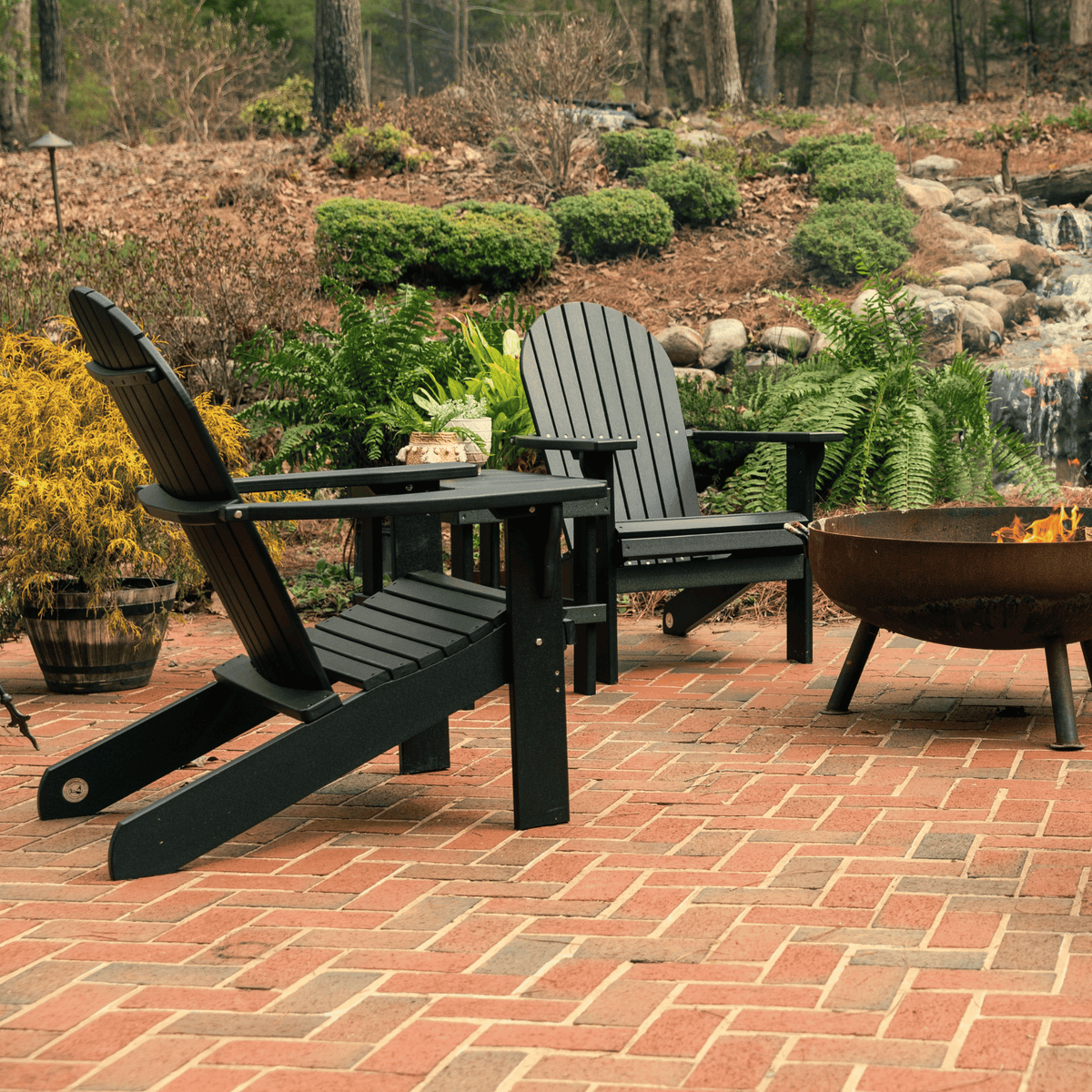 Two black Adirondack chairs on a brick patio with a fire pit in a garden setting.