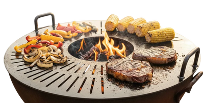 A round outdoor fire pit grill with handles sits on a gravel patio, cooking two steaks, corn on the cob, and sliced peppers and mushrooms over an open flame. Two patio chairs and stacked firewood are visible in the background of this backyard scene.