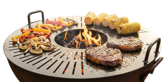 A round outdoor fire pit grill with handles sits on a gravel patio, cooking two steaks, corn on the cob, and sliced peppers and mushrooms over an open flame. Two patio chairs and stacked firewood are visible in the background of this backyard scene.
