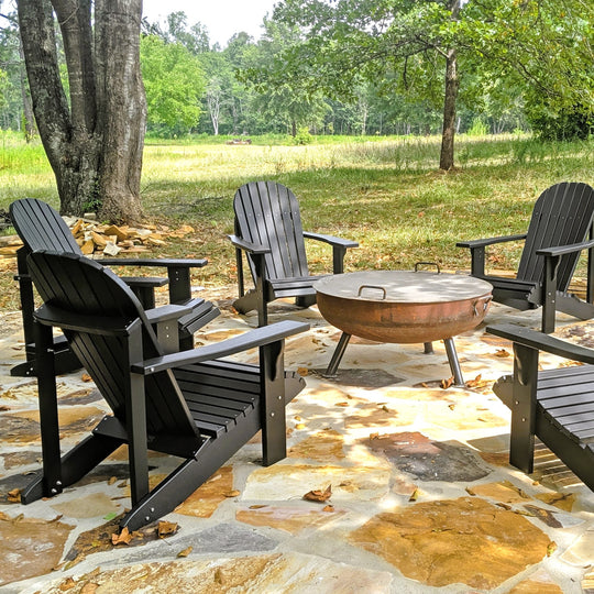 Black Adirondack chairs around a fire pit on a stone patio with trees in the background.